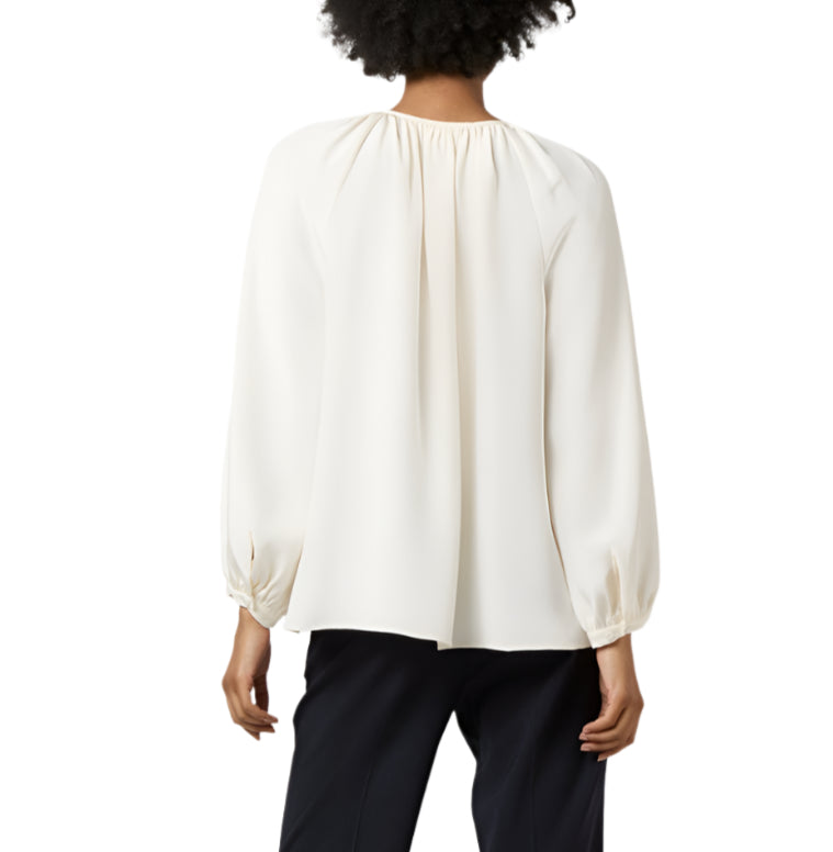 A woman is shown from the back against a plain white background, wearing the Ariana Blouse by Ann Mashburn—a loose-fitting, long-sleeved feminine top in white—paired with black pants.