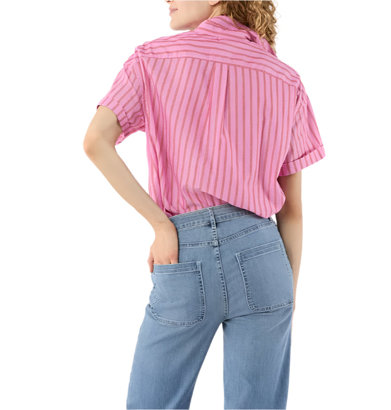 A person stands with one hand on their hip, wearing the Ann Mashburn Agnes Shirt—an oversized pink short-sleeve striped top—and light blue jeans, viewed from the back.