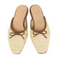 The Ann Mashburn Joy Basketweave Mule features beige slip-on flats with an almond-shaped toe, brown trim, and a small bow detail.