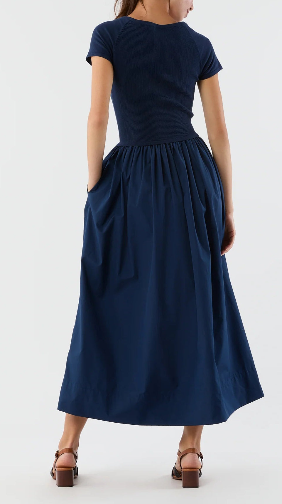 A woman faces away in the Ann Mashburn Cosette Cotton Silk Dress, featuring a navy short-sleeved fitted top and gathered poplin skirt, paired with brown heeled sandals.