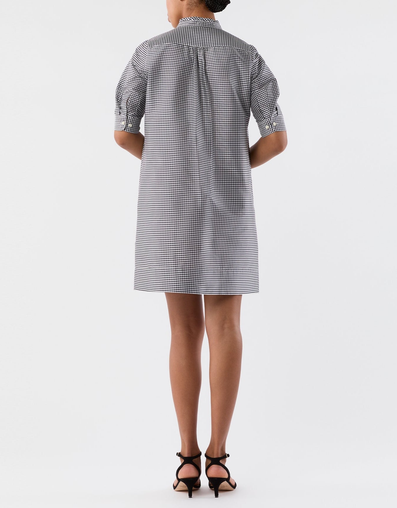A woman faces away, wearing the Ann Mashburn Elbow Sleeve Frill Dress—a feminine black and white checkered short dress with elbow-length sleeves—paired with black heeled sandals against a plain white background.