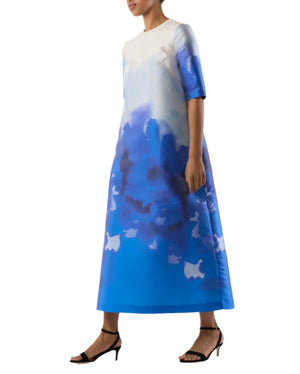 Wearing the Ann Mashburn Paige Maxi Dress in a blue and white gradient with short sleeves and black heeled sandals, someone stands against a plain background—perfect attire for a summer wedding.