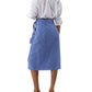 A person stands with their back to the camera, wearing a white long-sleeve shirt tucked into the Kara Wrap Skirt by Ann Mashburn, featuring blue and white stretch jacquard, a tie belt, and nude heeled sandals.