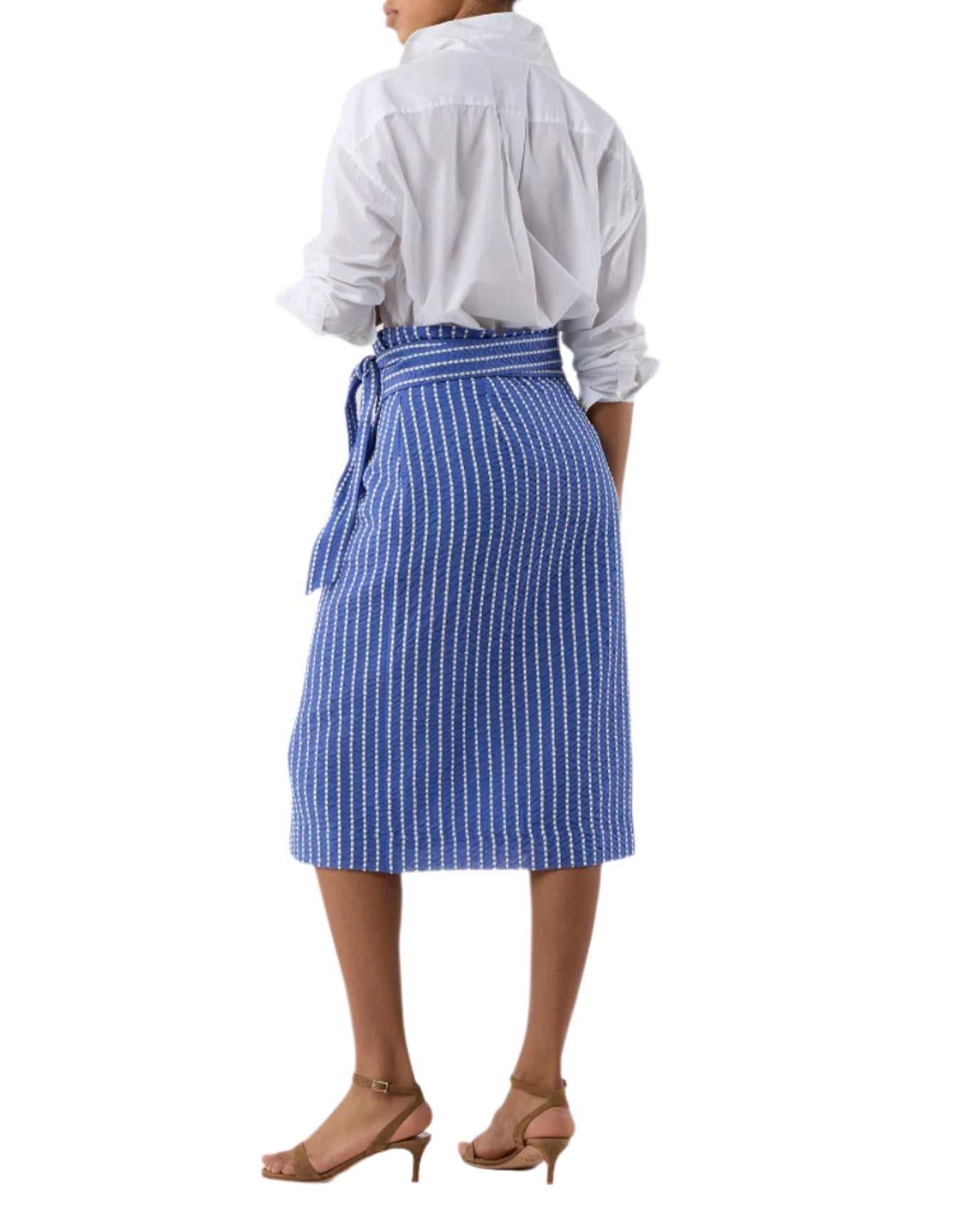 A person stands with their back to the camera, wearing a white long-sleeve shirt tucked into the Kara Wrap Skirt by Ann Mashburn, featuring blue and white stretch jacquard, a tie belt, and nude heeled sandals.