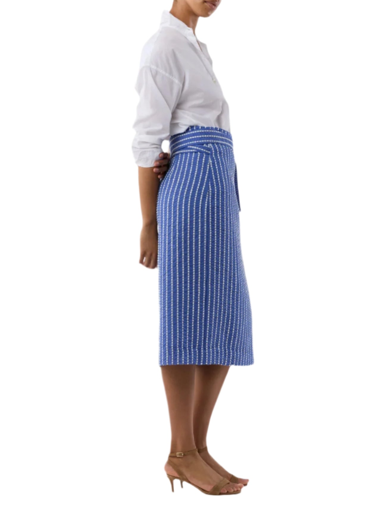 A woman stands in profile wearing a white long-sleeve shirt, tan heeled sandals, and the Kara Wrap Skirt by Ann Mashburn—a high-waisted skirt with blue and white vertical stripes.