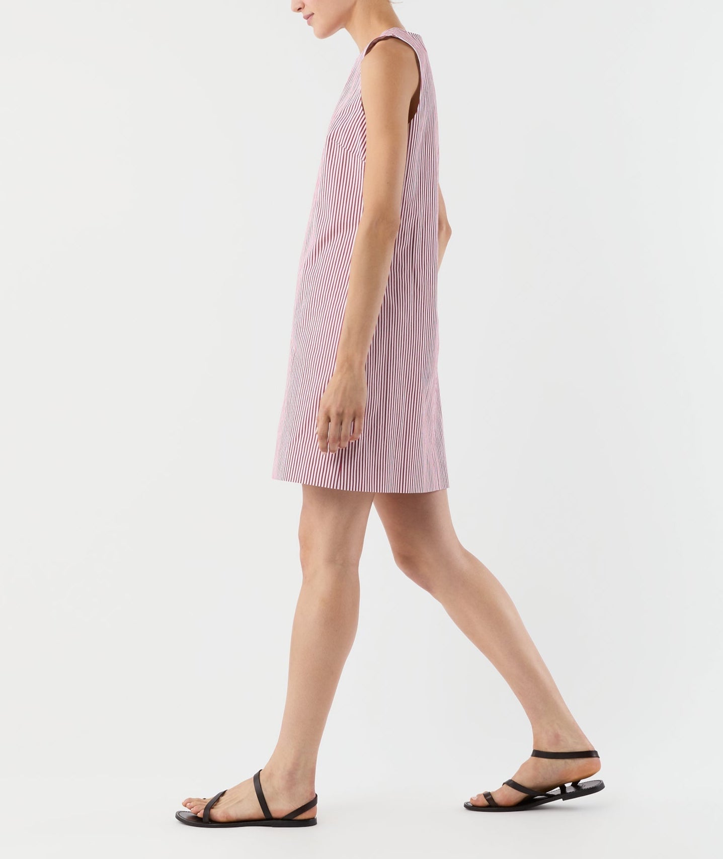 A person is walking against a plain white background wearing the Ann Mashburn Susy Shift Dress—a sleeveless, red-and-white striped 1960s A-line piece—paired with black sandals.
