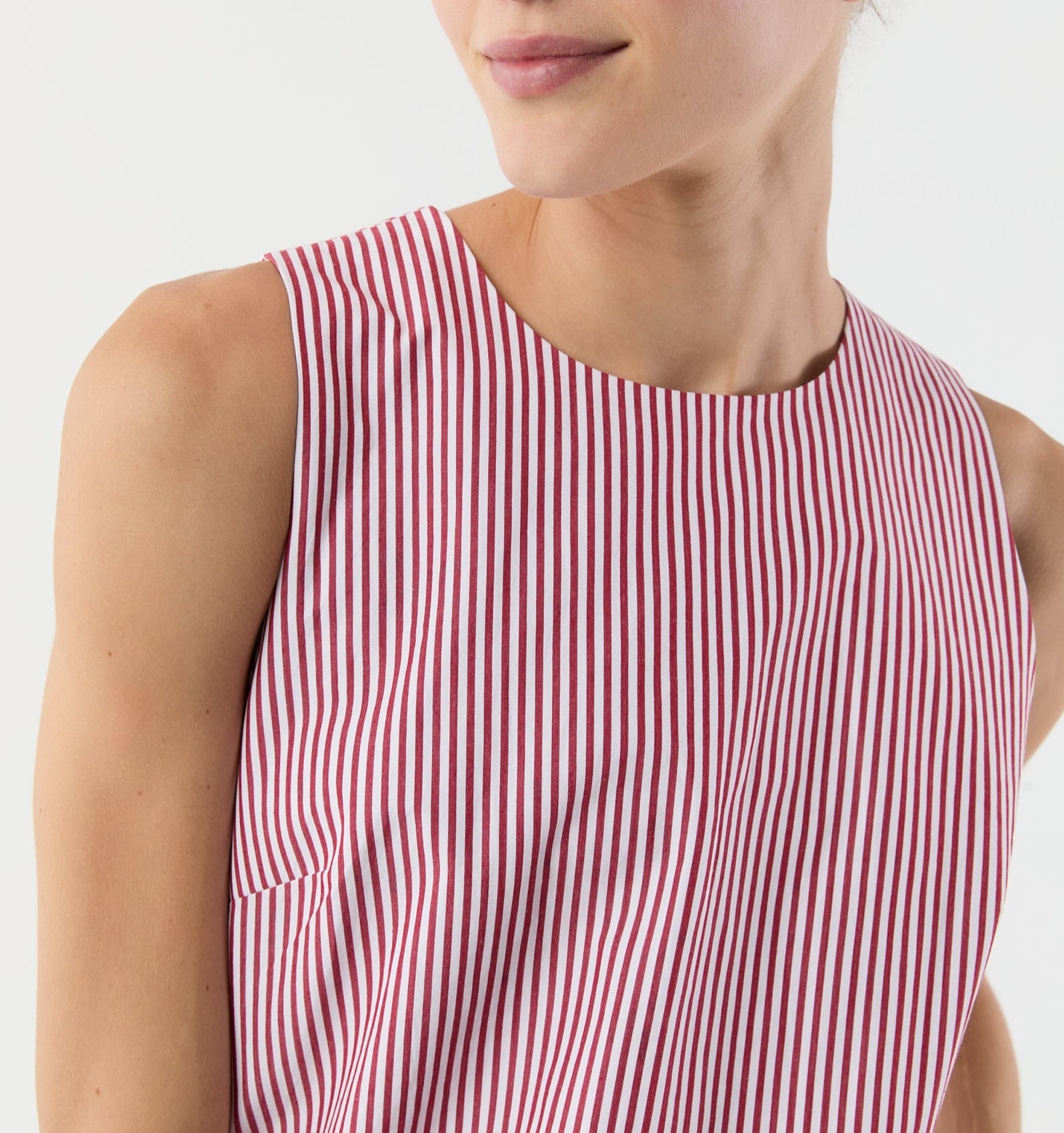 A person is shown from the shoulders up, facing slightly to the right, wearing the Susy Shift Dress by Ann Mashburn—a sleeveless dress with vertical red and white stripes made from Japanese compact cotton.