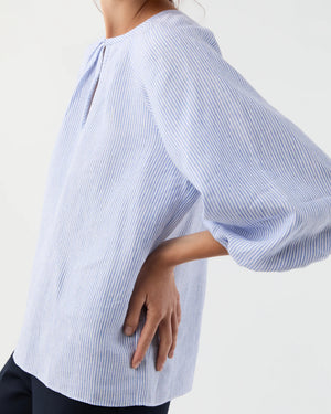 A person wears the Ann Mashburn Victoria Small Bengal Stripe Linen Blouse—light blue and white stripes, Belgian linen, keyhole neckline—perfect for spring and summer, paired with dark pants and posing with one hand on their hip.