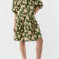 A person wears the Ann Mashburn Allegra Dress, featuring a green and beige floral seersucker fabric with puff sleeves and a tiered skirt, paired with brown heeled sandals, against a plain background.