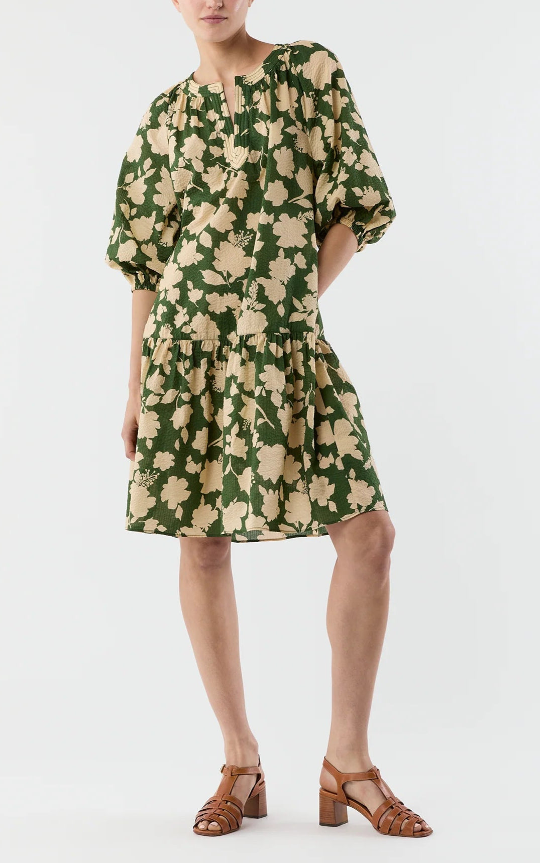 A person wears the Ann Mashburn Allegra Dress, featuring a green and beige floral seersucker fabric with puff sleeves and a tiered skirt, paired with brown heeled sandals, against a plain background.