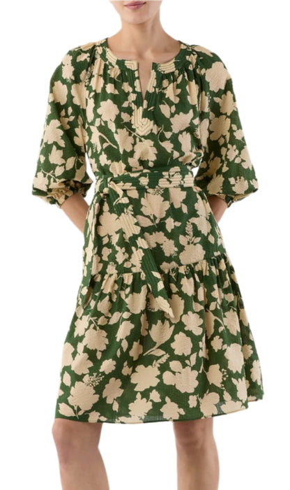 A woman wears the Ann Mashburn Allegra Dress, a green seersucker style with beige floral patterns, three-quarter sleeves, a belted waist, and a tiered skirt.