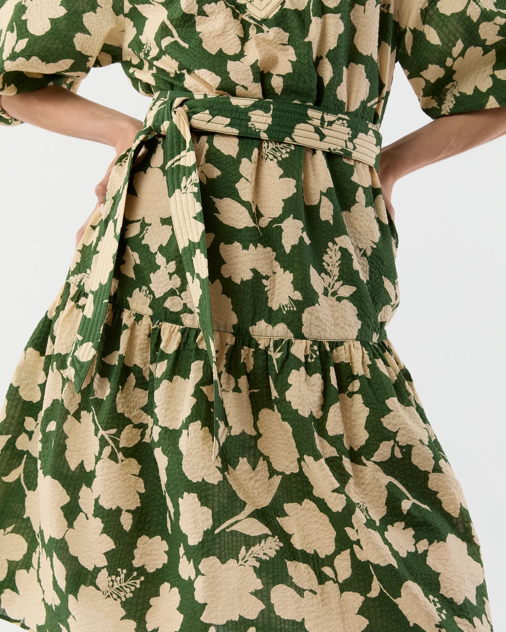 A close-up of the Allegra Dress by Ann Mashburn, a green seersucker style with beige floral prints, a tied waist belt, and a ruffled tiered skirt.