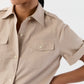 A person in the Ann Mashburn Short Sleeved Poplin Safari Shirt, beige with front pockets, stands against a plain light background.