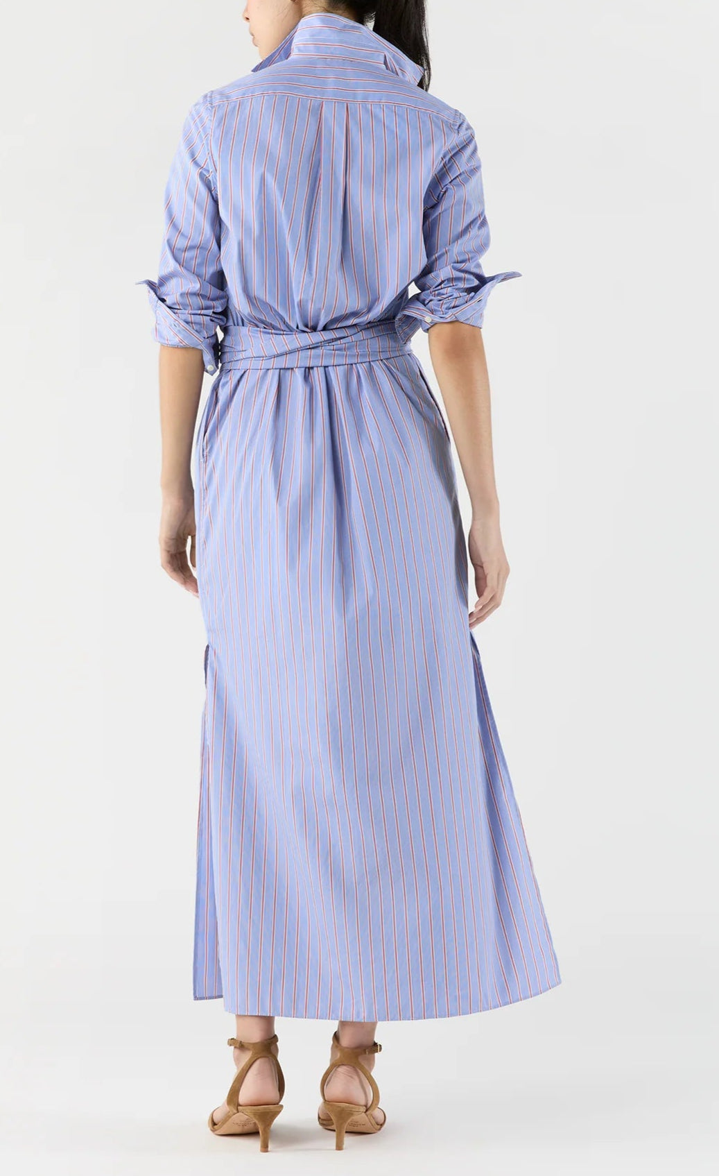 A person faces away, showcasing the back of the Ann Mashburn Maxi Tie-Waist Shirtdress in blue striped Italian poplin with long sleeves and a waist tie, paired with tan heeled sandals.