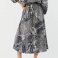A person wears the Ann Mashburn Miranda Block Print Skirt—a long black and white patterned midi skirt with an elastic waist—paired with a matching top and brown heeled sandals, standing against a plain background.
