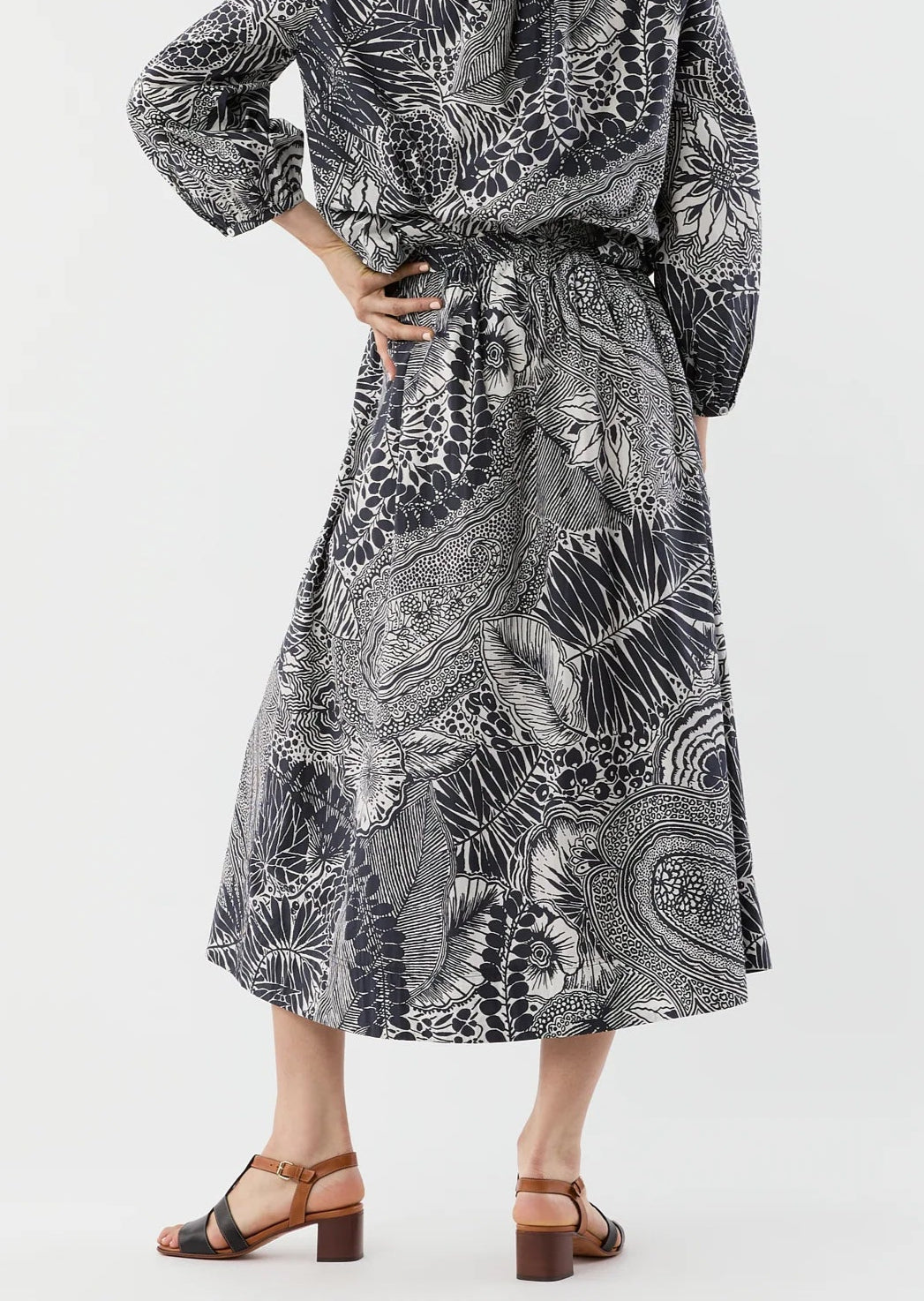 A person wears the Ann Mashburn Miranda Block Print Skirt—a long black and white patterned midi skirt with an elastic waist—paired with a matching top and brown heeled sandals, standing against a plain background.