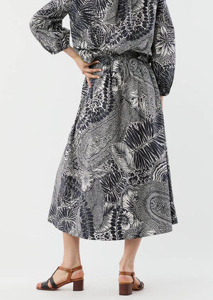 A person wears the Ann Mashburn Miranda Block Print Skirt—a long black and white patterned midi skirt with an elastic waist—paired with a matching top and brown heeled sandals, standing against a plain background.