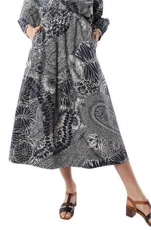 A person wears the Ann Mashburn Miranda Block Print Skirt, a black and white patterned midi with an elastic waist and pockets, paired with black heeled sandals, standing against a plain background.