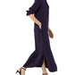 A person wearing the Ann Mashburn Piper Maxi Dress in dark purple crepe de chine with long sleeves, side slits, and black heeled sandals walks against a white background.