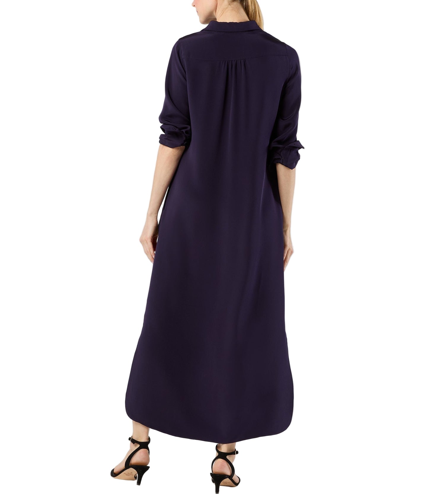 A woman stands with her back to the camera, wearing an ankle-length, lightweight dark purple Piper Maxi Dress by Ann Mashburn with long sleeves and black heeled sandals.