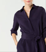 A woman stands against a plain light background wearing the Ann Mashburn Piper Maxi Dress in dark purple crepe de chine, featuring a waist tie and partially rolled-up sleeves.