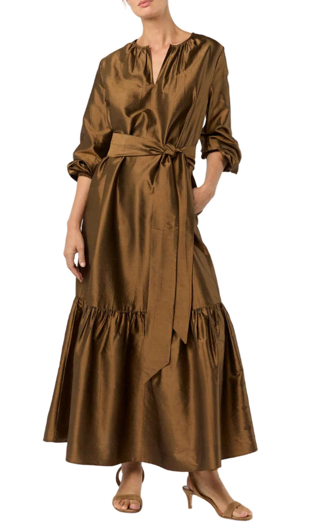 A person wearing the Aba Dress by Ann Mashburn—a long copper-brown silk shantung maxidress with a self-tie belt, long sleeves, and ruffled hem—paired with tan open-toe heels.
