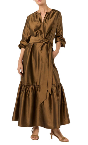 A person wearing the Aba Dress by Ann Mashburn—a long copper-brown silk shantung maxidress with a self-tie belt, long sleeves, and ruffled hem—paired with tan open-toe heels.