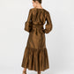 A woman faces away in the Ann Mashburn Aba Dress, a bronze silk shantung maxidress with long sleeves, gathered skirt, self-tie waist belt, and heeled sandals. Her hair is styled in a neat bun.