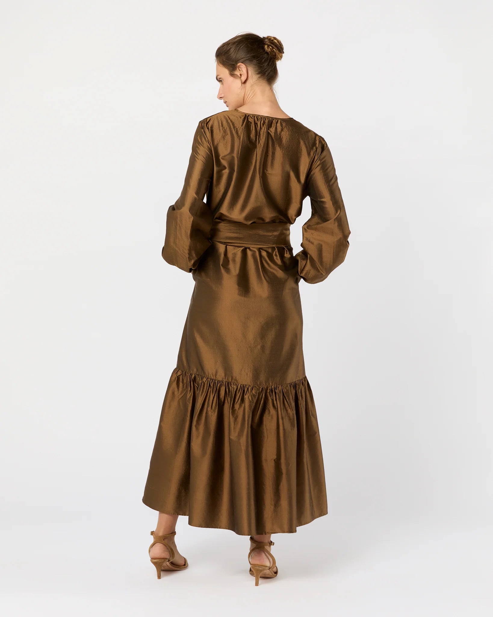 A woman faces away in the Ann Mashburn Aba Dress, a bronze silk shantung maxidress with long sleeves, gathered skirt, self-tie waist belt, and heeled sandals. Her hair is styled in a neat bun.
