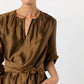 A woman wears the Ann Mashburn Aba Dress, a shiny brown silk shantung style with a gathered neckline and self-tie belt, posing with one hand in her pocket against a plain background.