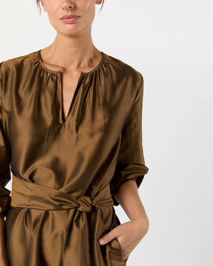 A woman wears the Ann Mashburn Aba Dress, a shiny brown silk shantung style with a gathered neckline and self-tie belt, posing with one hand in her pocket against a plain background.