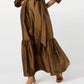 A person wearing the Ann Mashburn Aba Dress—a long brown silk maxi with a self-tie belt, puff sleeves, and ruffled hem—paired with tan heeled sandals.