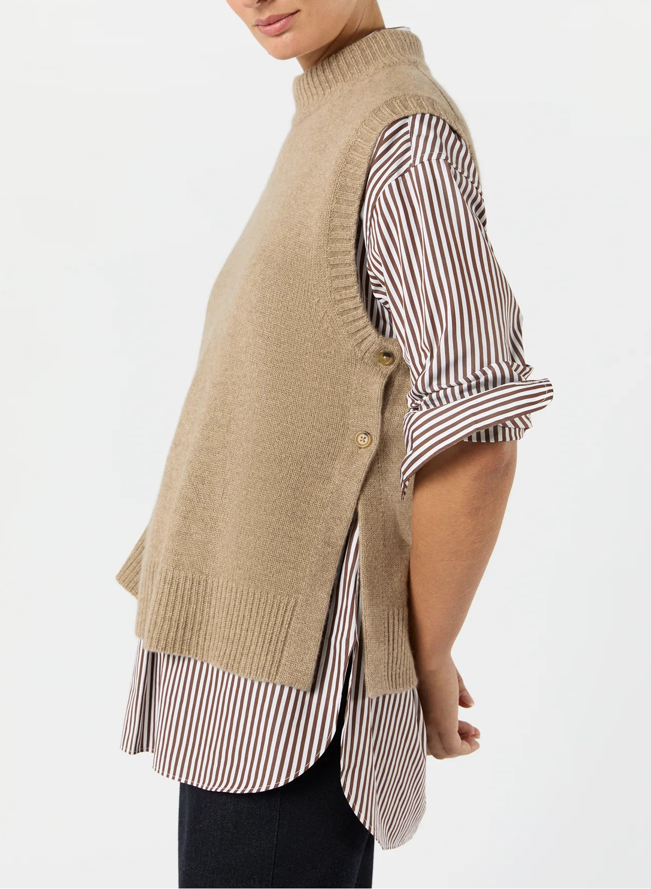 A person models the Ann Mashburn Marion Sweater, a beige cashmere vest, layered over a brown-and-white striped long-sleeve shirt with black pants, shown in profile against a plain background.