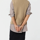 A person stands facing away, wearing the Ann Mashburn Marion Sweater vest in tan over a striped shirt and dark skirt, hands clasped behind their back.