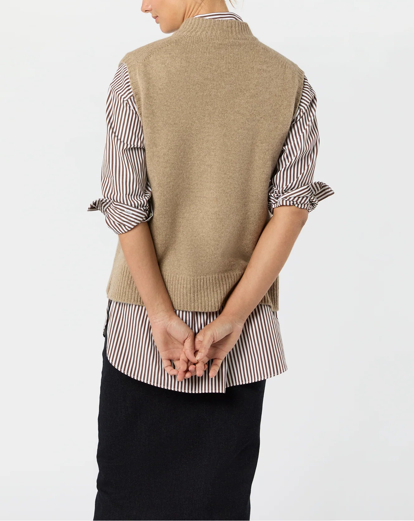 A person stands facing away, wearing the Ann Mashburn Marion Sweater vest in tan over a striped shirt and dark skirt, hands clasped behind their back.