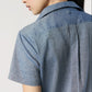 A person with long dark hair is seen from behind in an Ann Mashburn Scout Cotolino Chambray Shirt, short-sleeved and light blue, against a plain light background.