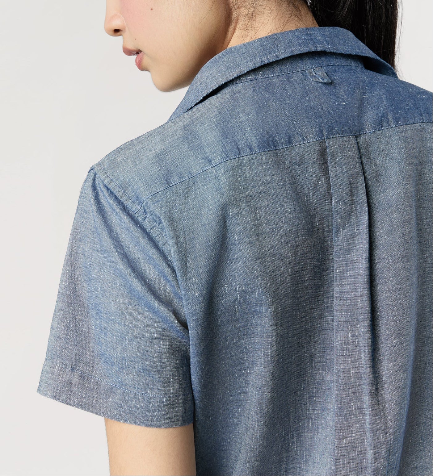 A person with long dark hair is seen from behind in an Ann Mashburn Scout Cotolino Chambray Shirt, short-sleeved and light blue, against a plain light background.