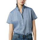 A person wearing a short-sleeve, light blue Ann Mashburn Scout Cotolino Chambray Shirt with a popped collar and blue jeans, shown from shoulders to hips.