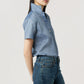 A person wears the Ann Mashburn Scout Cotolino Chambray Shirt with short sleeves and collar turned up, paired with blue jeans, standing against a plain light background.
