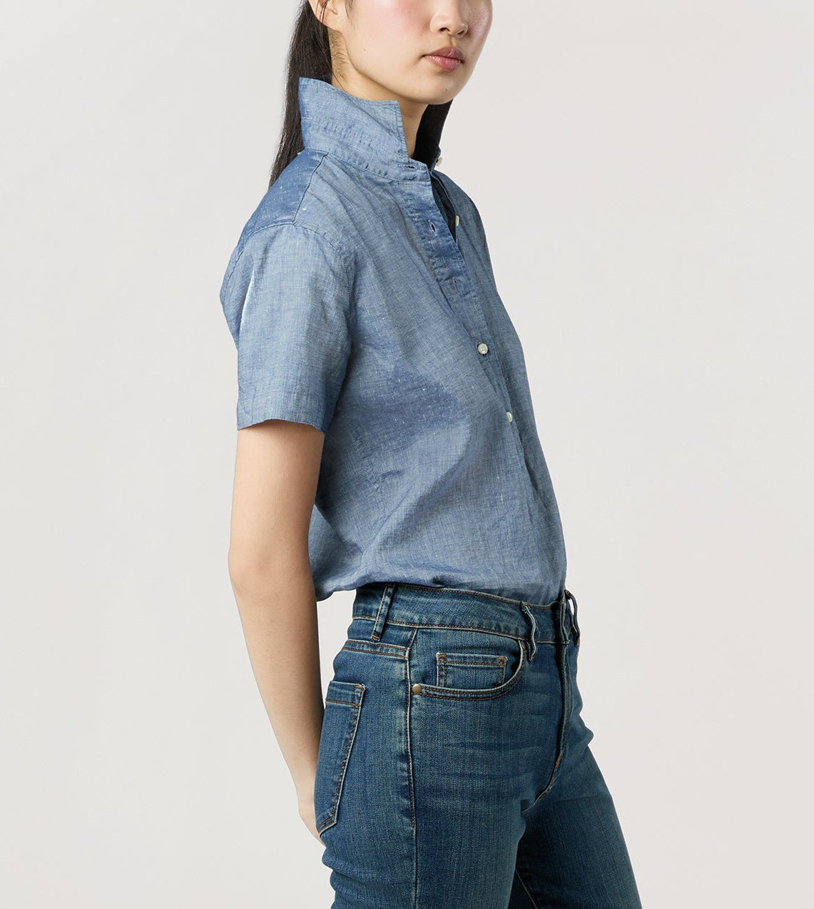 A person wears the Ann Mashburn Scout Cotolino Chambray Shirt with short sleeves and collar turned up, paired with blue jeans, standing against a plain light background.