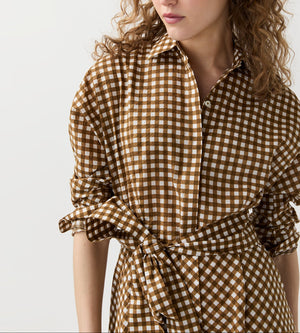 A woman wears the Ann Mashburn Charlie Shirtwaist Gingham Dress in brown and white seersucker, featuring rolled-up sleeves and a tied waist, standing against a plain light background.