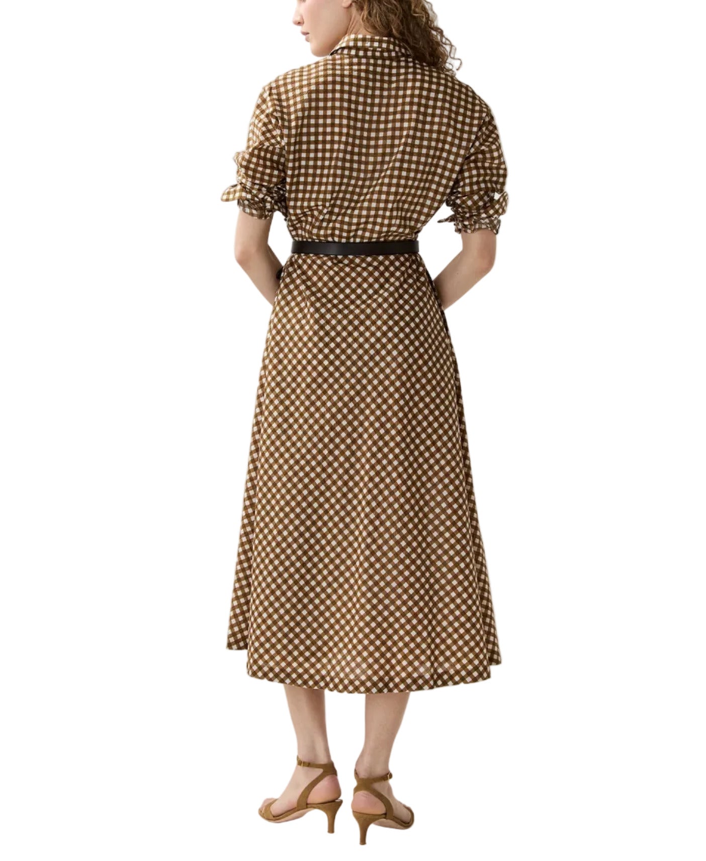 A woman, viewed from the back, stands in heeled sandals wearing the Ann Mashburn Charlie Shirtwaist Gingham Dress—a brown and white checkered midi shirtdress with short sleeves and a black belt.