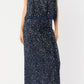A woman stands against a plain background wearing the Ann Mashburn Gia Beaded Mesh Shell, a sleeveless dark blue sequined top, paired with black block-heeled sandals.