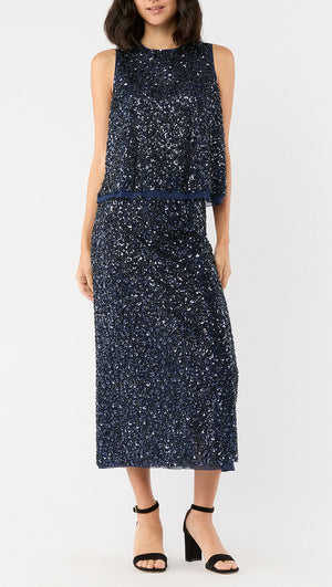 A woman stands against a plain background wearing the Ann Mashburn Gia Beaded Mesh Shell, a sleeveless dark blue sequined top, paired with black block-heeled sandals.