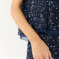 A person is shown from shoulder to thigh, wearing the Ann Mashburn Gia Beaded Mesh Shell in dark blue, with their left arm relaxed at their side against a plain background.