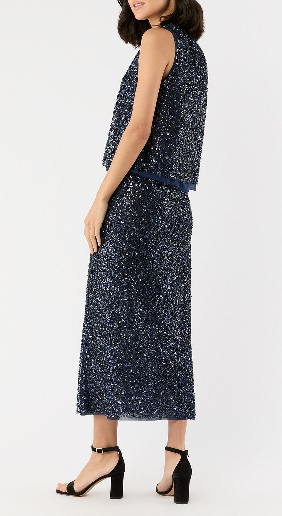 A woman stands against a white background wearing Ann Mashburn's Ruby Beaded Mesh Skirt paired with a sleeveless navy sequin top and black ankle-strap block heel sandals.