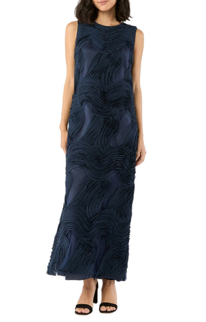 A woman wearing the Ann Mashburn Susy Shift Maxi Dress, a sleeveless, floor-length navy dress with textured wave patterns, paired with black heeled sandals.