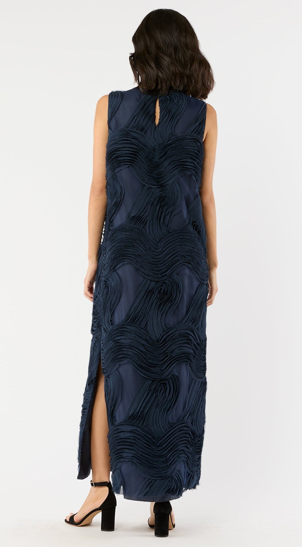 A woman is seen from the back against a white background wearing the Ann Mashburn Susy Shift Maxi Dress in sleeveless navy textured fabric with a side slit, paired with black block-heel sandals.
