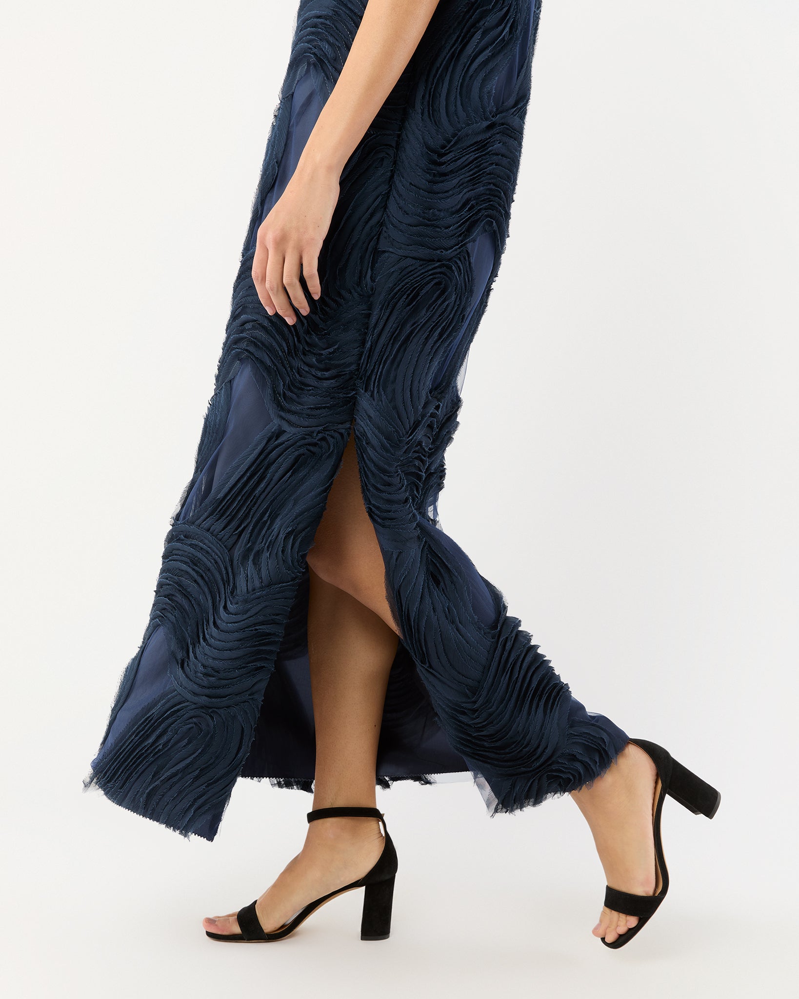 A person wearing the Ann Mashburn Susy Shift Maxi Dress in textured navy blue with a slit and black block-heeled sandals, walking against a plain light background.