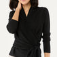 A woman wearing the Ann Mashburn Mika Cashmere Wrap Sweater, a luxury black long sleeve top with a tied waistband, stands with one hand touching her neck.